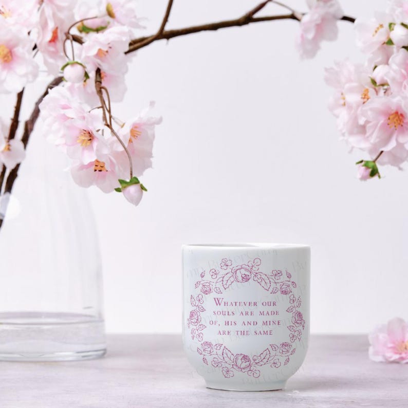 Puede incluir: Un vaso de cer&aacute;mica blanco con un dise&ntilde;o floral y la cita, "Whatever our souls are made of, his and mine are the same." Flores de cerezo rosas y un jarr&oacute;n transparente en el fondo.