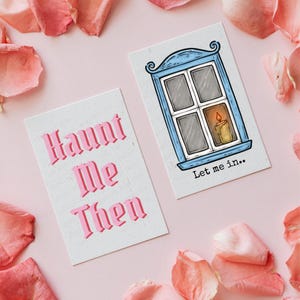 Puede incluir: Dos tarjetas rectangulares sobre un fondo rosa con p&eacute;talos de rosa. Una tarjeta dice "Haunt Me Then" en letras g&oacute;ticas rosas. La otra presenta una ilustraci&oacute;n de una ventana azul con una vela encendida y el texto "Let me in.."