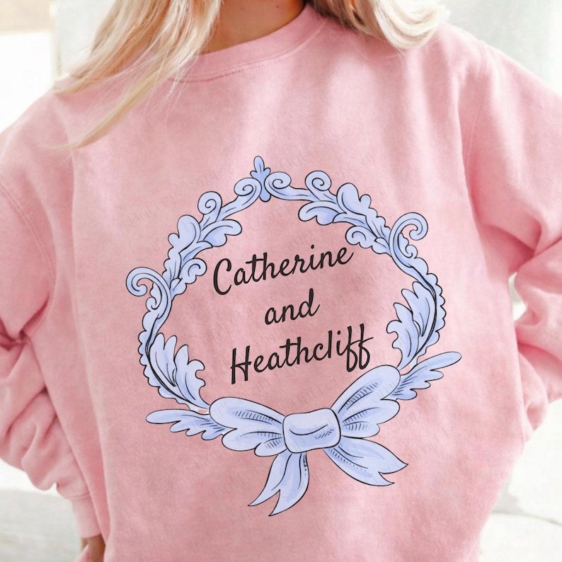 Puede incluir: Sudadera rosa claro con el texto "Catherine and Heathcliff" dentro de un marco azul claro adornado con un lazo. La sudadera es de color liso.