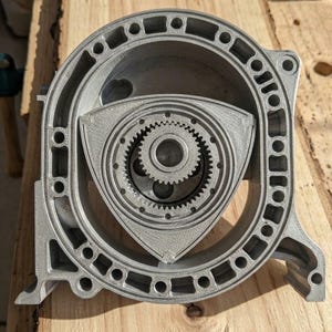 May include: A detailed, gray rotary engine model, showcasing the internal components. The model features a triangular rotor within a circular housing, with visible gears and intricate design elements. The engine is set against a wooden surface.
