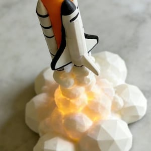 May include: A toy space shuttle, white and black with an orange nose, is shown taking off. It sits on a white, geometric cloud base with a warm, glowing light. A decorative item, possibly a night light, with a space theme.