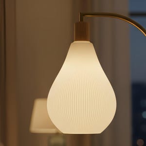 Puede incluir: Una lámpara moderna en forma de lágrima, con una pantalla de vidrio blanco acanalado. La lámpara presenta una base de madera y un brazo curvo de color latón. El brillo suave sugiere que está encendida, iluminando una habitación. Un elemento de decoración del hogar elegante.