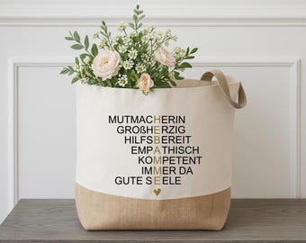 Könnte beinhalten: Eine weiße Canvas-Tragetasche mit braunem Jute-Besatz. Die Tasche hat goldene Schriftzüge mit der Aufschrift "MUTMACHERIN, GROSHERZIG, HILFSBEREIT, EMPATHISCH, KOMPETENT, IMMER DA, GUTE SEELE". Ein kleines goldenes Herz befindet sich am Boden der Tasche.