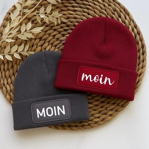 Könnte beinhalten: Zwei Strickmützen mit dem Wort "moin" auf der Vorderseite gestickt. Eine Mütze ist grau mit weißen Buchstaben und die andere ist rot mit weißen Buchstaben.