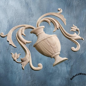 May include: A wooden decorative carving featuring a vase with flowing, stylized leaves and floral accents. The carving is made of light-colored wood and is set against a textured blue background. The design is symmetrical and detailed.