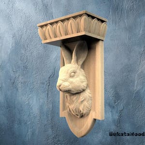 May include: A wooden wall shelf with a carved rabbit head. The shelf has a rectangular top with a decorative edge and a shield-shaped base. The rabbit head is intricately detailed, with visible fur and facial features. The shelf is made of light-coloured wood.