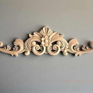Puede incluir: Elemento arquitectónico decorativo de madera con un diseño simétrico. La pieza presenta detalles florales y de volutas, con una flor central y acentos rizados. La madera es de color claro natural y está montada sobre un fondo gris.