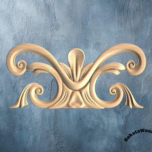 May include: A carved wooden decorative element with a symmetrical design. The piece features swirling scrollwork and a central floral motif. The wood has a natural, light brown color, and the background is a textured blue.
