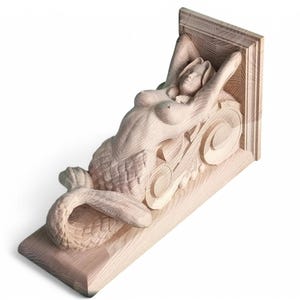 Pode incluir: Um corbel de madeira com uma figura de sereia esculpida. A sereia está deitada de lado com a cauda enrolada ao redor dela. O corbel é feito de madeira clara e tem um acabamento natural.