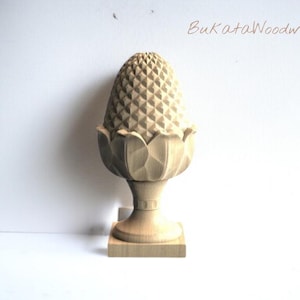 Op de afbeelding: Een gesneden houten ananas-sierstuk met een gedetailleerd, getextureerd oppervlak. Het sierstuk staat op een decoratieve basis met een bloemenontwerp en een vierkante sokkel. De tekst "BukataWoodwork" is rechtsboven zichtbaar.