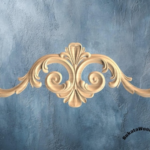 May include: A carved wooden decorative scroll with a symmetrical design. The scroll features a central floral element and curved, swirling details. The wood has a natural, light brown colour, and the background is a textured blue.