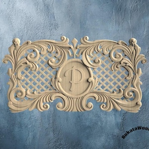 May include: A carved wooden decorative panel with an ornate design. The panel features scrolling floral elements, a lattice background, and a central medallion with the letter "P". The panel is light brown in color.