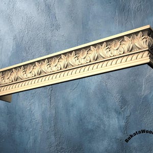Puede incluir: Una moldura arquitectónica de madera tallada con un diseño repetitivo de hojas y volutas. La madera de color claro está montada sobre una pared azul texturizada. La moldura es un elemento decorativo para el diseño de interiores.