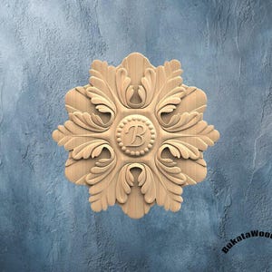Op de afbeelding: Een gesneden houten bloemenmedaillon met een centrale 'B' en ingewikkelde blad details. Het medaillon is lichtbruin en geplaatst tegen een getextureerde blauwe achtergrond. Het ontwerp is symmetrisch en gedetailleerd.