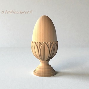 May include: A wooden egg-shaped sculpture in a decorative holder. The holder features a carved floral design. The sculpture is made of light-colored wood and stands on a pedestal base. The text "BuKataWoodwork" is in the upper left corner.