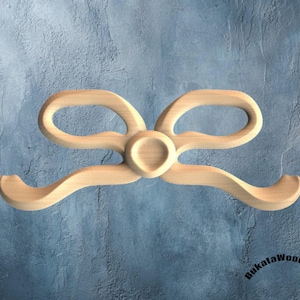 Könnte beinhalten: Ein dekoratives Element in Form einer Holzschleife. Der Bogen ist aus hellfarbenem Holz geschnitzt und weist zwei Schlaufen und gebogene Enden auf. Das Design umfasst ein zentrales kreisförmiges Detail. Der Hintergrund ist eine strukturierte blaue Oberfläche.
