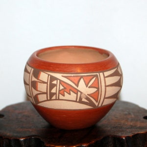 Può includere: Una piccola ciotola in terracotta dipinta a mano con un motivo geometrico in tonalità di marrone, arancio e bianco. La ciotola è appoggiata su una base in legno.