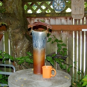 May include: A brown and blue ceramic vase with a unique handle sits on a gray table next to a small orange mug. The vase is decorated with a drip glaze pattern. The table is outdoors, with a wooden fence and green plants in the background.