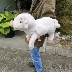 May include: A plush pig toy with soft, light pink fur, a snout, floppy ears, and small tan hooves. The stuffed animal has a curled tail and a small white tag attached to its side, showcasing a realistic design with a rounded body and four legs.