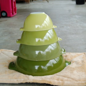 May include: A stack of four green mixing bowls, nested together, with a white rim on the top bowl. The bowls are sitting on a beige, fuzzy mat.