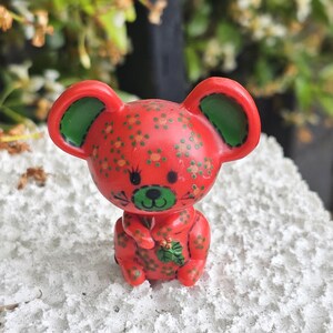 May include: A small red mouse figurine featuring a pattern of tiny orange and green flowers. The mouse has large ears with green interiors, a green snout, black eyes, and holds a small green holly leaf sprig on its chest area.
