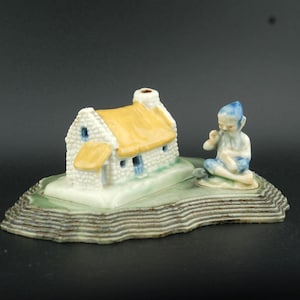 May include: A ceramic figurine depicting a small cottage with a yellow thatched roof and a seated figure. The cottage is white with blue accents, and the base is green and brown.