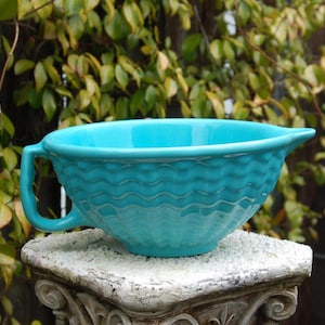 May include: A turquoise ceramic mixing bowl with a handle and a spout. The bowl has a textured, wavy pattern around the exterior. The bowl is sitting on a white, ornate pedestal, with green foliage in the background.