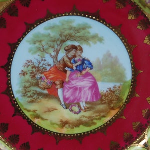 Peut inclure: Une assiette en porcelaine rouge et or avec une image centrale d'un couple dans un cadre romantique. Le couple est entour&eacute; d'arbres et de fleurs. L'assiette a un bord dor&eacute; et un motif dor&eacute; autour du bord.