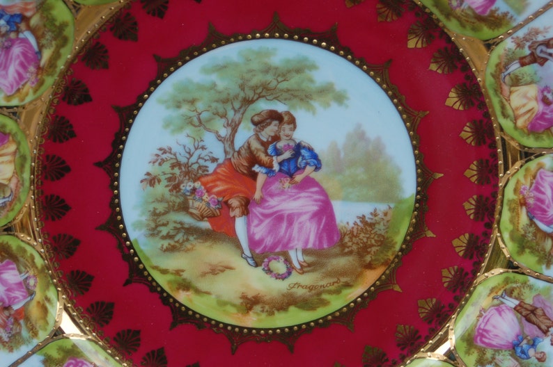 Peut inclure: Une assiette en porcelaine rouge et or avec une image centrale d'un couple dans un cadre romantique. Le couple est entour&eacute; d'arbres et de fleurs. L'assiette a un bord dor&eacute; et un motif dor&eacute; autour du bord.