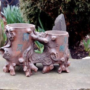 Puede incluir: Un macetero de cerámica marrón con forma de tronco de árbol con dos macetas. Las macetas tienen caracteres chinos. El macetero está sobre una superficie de piedra gris.