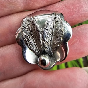 May include: A silver-toned metal brooch shaped like a flower with five smooth, rounded petals. Two textured, veined leaves are positioned in the center, and a small, polished spherical bead sits at the base of the leaves, completing the floral design.