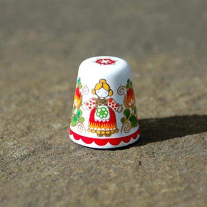 Könnte beinhalten: Weiße Keramik-Fingerhut mit einem roten, weißen und grünen Blumendesign, das ein Mädchen in einem roten Kleid zeigt.