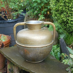 Puede incluir: Una jarra de latón antigua grande con dos asas. La jarra tiene una pátina desgastada y envejecida y una banda decorativa alrededor del centro.