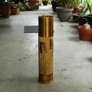 May include: A tall, cylindrical brass vase with a textured, etched design of reeds or tall grass. The top and bottom are smooth and shiny, while the middle section has a rougher, more detailed appearance. The vase is standing on a concrete surface.