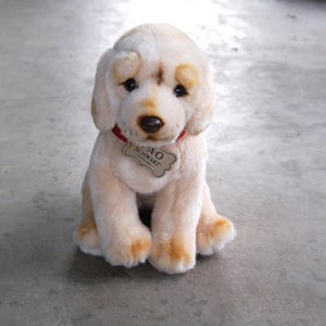 May include: A plush golden retriever puppy with a red collar and a tag that reads "A.O. Schwarz". The stuffed animal has brown accents on its ears, nose, and paws. It is sitting on a gray surface.