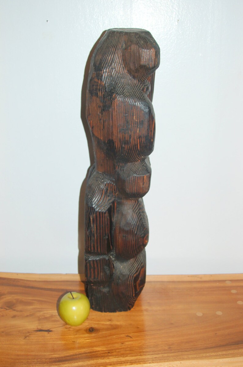 Op de afbeelding: Een donkerbruin houten beeldhouwwerk van een gestileerde menselijke figuur. Het beeldhouwwerk is gebeeldhouwd met een ruw, getextureerd oppervlak en heeft een afgeronde, abstracte vorm.