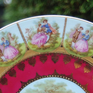 Peut inclure: Une assiette en porcelaine vintage avec un bord rouge et or. L'assiette pr&eacute;sente un motif central d'un couple dans un jardin, entour&eacute; de huit sc&egrave;nes plus petites de couples dans diverses poses romantiques.
