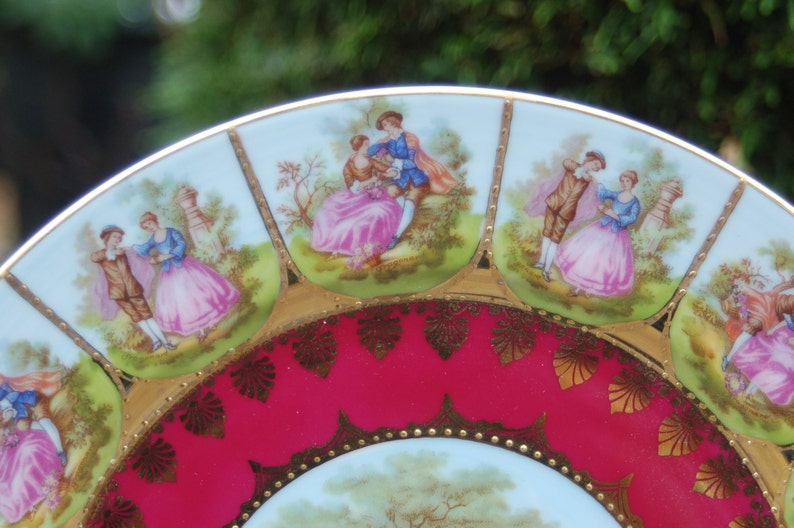 Peut inclure: Une assiette en porcelaine vintage avec un bord rouge et or. L'assiette pr&eacute;sente un motif central d'un couple dans un jardin, entour&eacute; de huit sc&egrave;nes plus petites de couples dans diverses poses romantiques.