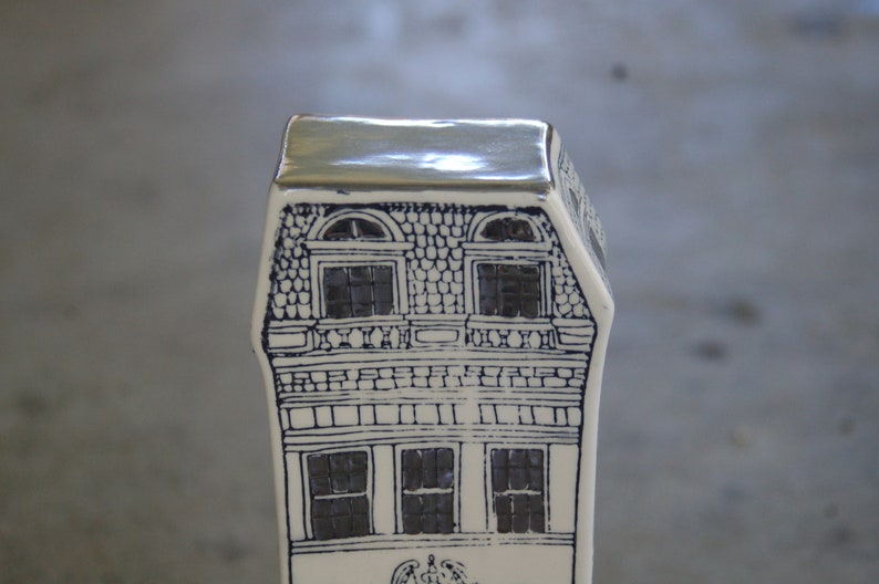 May include: A white ceramic house figurine with a silver roof and black and white details. The house has two windows on the top floor and three windows on the bottom floor.