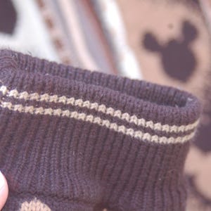 Peut inclure: Gros plan sur un poignet de chaussette en tricot marron fonc&eacute; avec deux rang&eacute;es de coutures beige clair. Le poignet est c&ocirc;tel&eacute; et a une texture douce. L'arri&egrave;re-plan est flou, montrant un motif avec des silhouettes de Mickey Mouse.