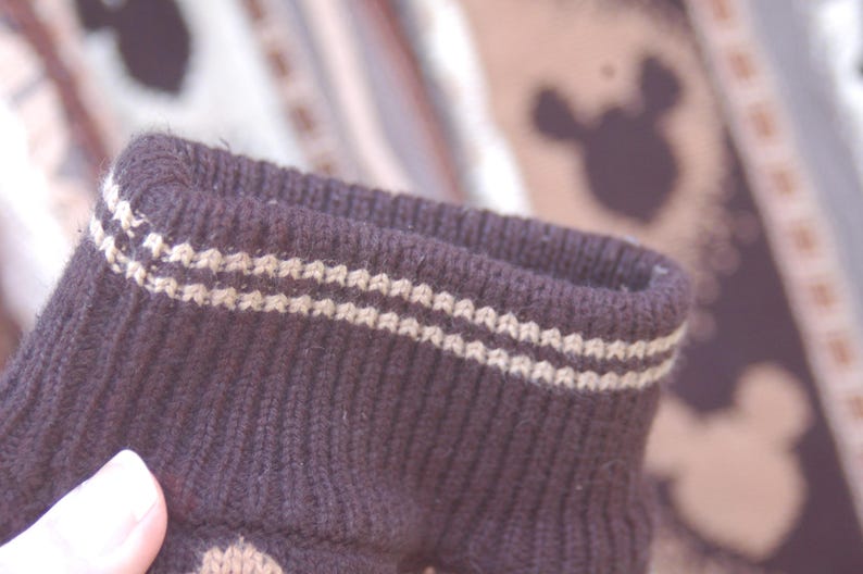 Peut inclure: Gros plan sur un poignet de chaussette en tricot marron fonc&eacute; avec deux rang&eacute;es de coutures beige clair. Le poignet est c&ocirc;tel&eacute; et a une texture douce. L'arri&egrave;re-plan est flou, montrant un motif avec des silhouettes de Mickey Mouse.