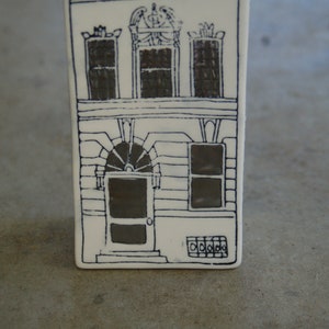 May include: A white ceramic tile with a black line drawing of a house with three windows and a door. The house has a decorative design above the windows.
