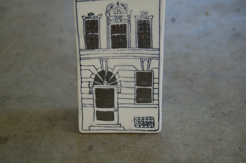 May include: A white ceramic tile with a black line drawing of a house with three windows and a door. The house has a decorative design above the windows.