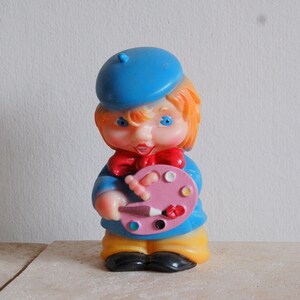 Op de afbeelding: Een blauw en geel plastic speelgoedfiguur van een schilder die een blauwe baret draagt en een roze palet met een penseel vasthoudt.