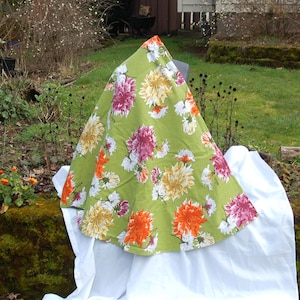 Könnte beinhalten: Ein grüner Stoff mit einem lebendigen Blumenmuster in Orange, Rosa, Gelb und Weiß. Der Stoff ist drapiert und zeigt das Design. Der Hintergrund ist eine natürliche Außenumgebung mit Grün und einem weißen Tuch.