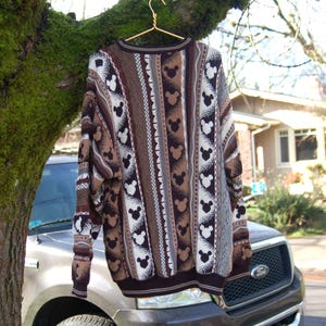 Peut inclure: Un pull en tricot marron et blanc avec un motif Mickey Mouse. Le pull a un col rond et des manches longues. Le motif comprend des rayures verticales et des silhouettes de Mickey Mouse. Il est suspendu &agrave; un cintre dor&eacute;.