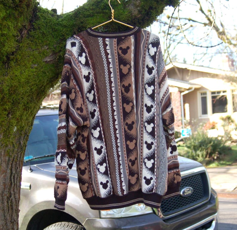 Peut inclure: Un pull en tricot marron et blanc avec un motif Mickey Mouse. Le pull a un col rond et des manches longues. Le motif comprend des rayures verticales et des silhouettes de Mickey Mouse. Il est suspendu &agrave; un cintre dor&eacute;.