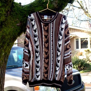 Peut inclure: Un pull &agrave; manches longues orn&eacute; de motifs Mickey Mouse noirs et blancs, alternant avec des rayures marron et beige. Le pull est suspendu &agrave; un cintre dor&eacute; sur une branche d'arbre.