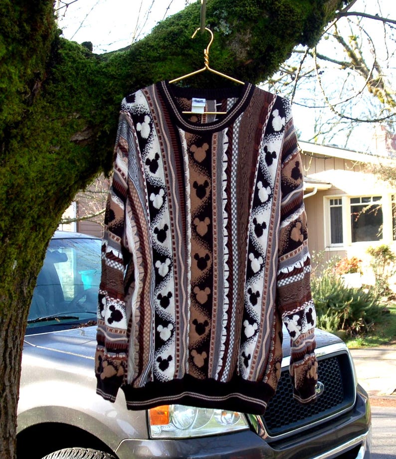 Peut inclure: Un pull &agrave; manches longues orn&eacute; de motifs Mickey Mouse noirs et blancs, alternant avec des rayures marron et beige. Le pull est suspendu &agrave; un cintre dor&eacute; sur une branche d'arbre.