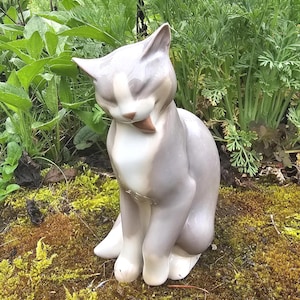 Könnte beinhalten: Eine sitzende Keramik-Katzenfigur mit geschlossenen Augen und leicht geöffnetem Mund. Die Katze ist überwiegend grau mit weißen Akzenten und einer braunen Zunge. Sie befindet sich im Freien zwischen grünen Pflanzen und Moos.